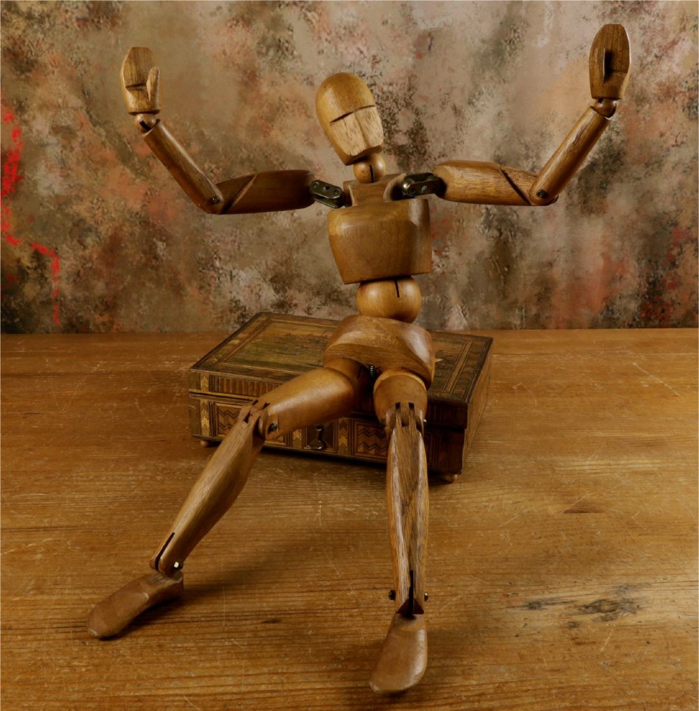 A carved teak articulated artist lay figure standing on a surface with both arms raised at the elbow joints. View II
