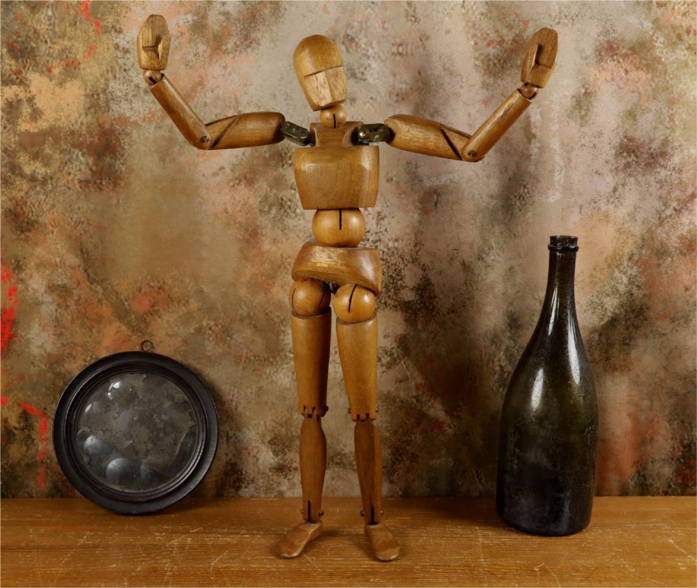 A carved teak articulated artist lay figure standing on a surface with both arms raised at the elbow joints. View IV