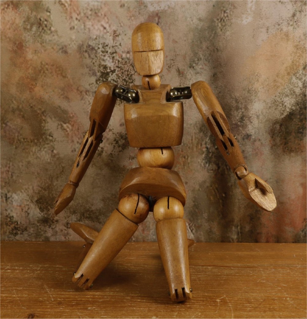 A carved teak articulated artist lay figure standing on a surface with both arms raised at the elbow joints. View VI