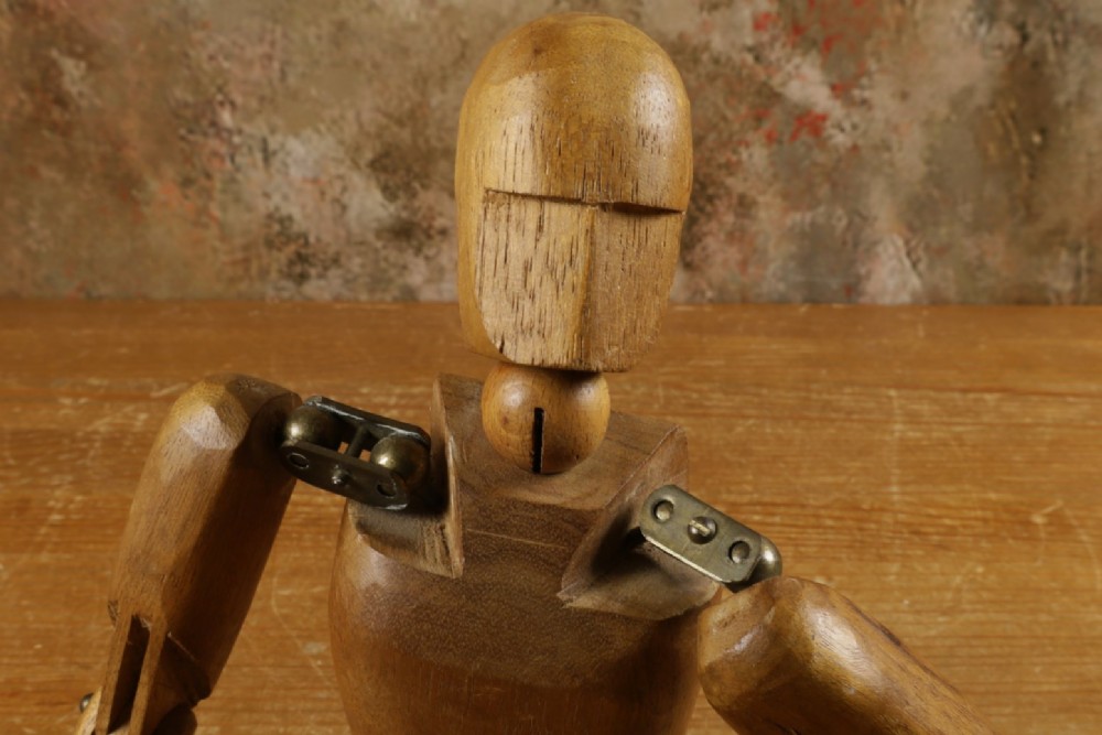 A carved teak articulated artist lay figure standing on a surface with both arms raised at the elbow joints. View VII