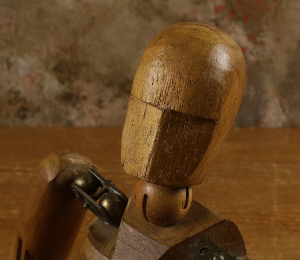 A carved teak articulated artist lay figure standing on a surface with both arms raised at the elbow joints. View VIII