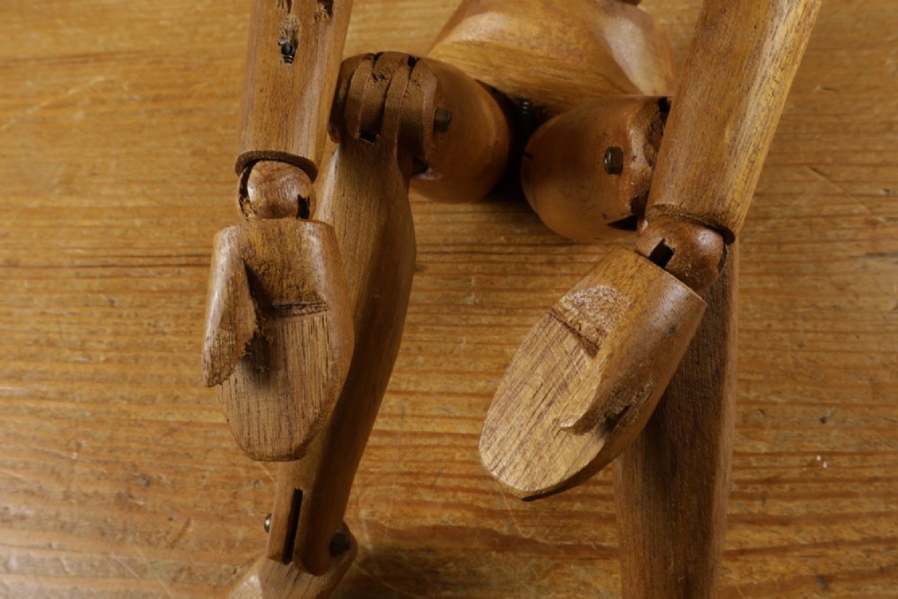 A carved teak articulated artist lay figure standing on a surface with both arms raised at the elbow joints. View X