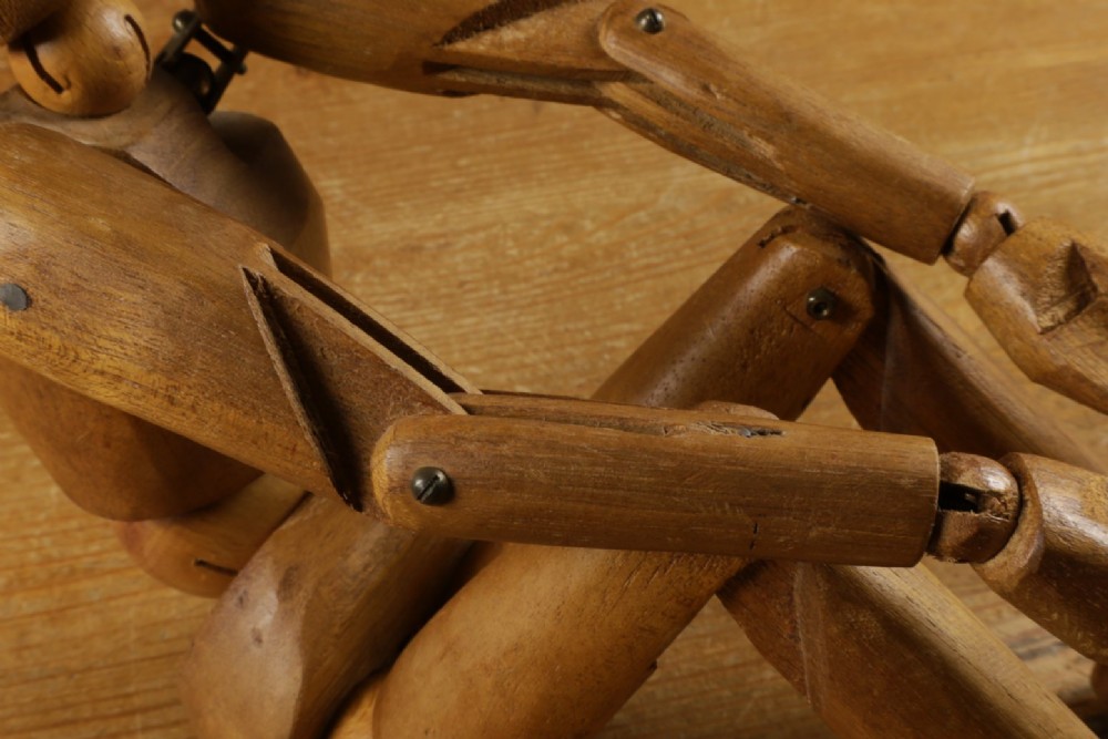 A carved teak articulated artist lay figure standing on a surface with both arms raised at the elbow joints. View XI
