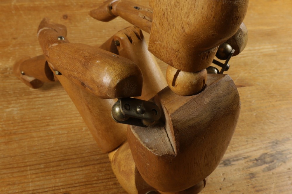 A carved teak articulated artist lay figure standing on a surface with both arms raised at the elbow joints. View XV