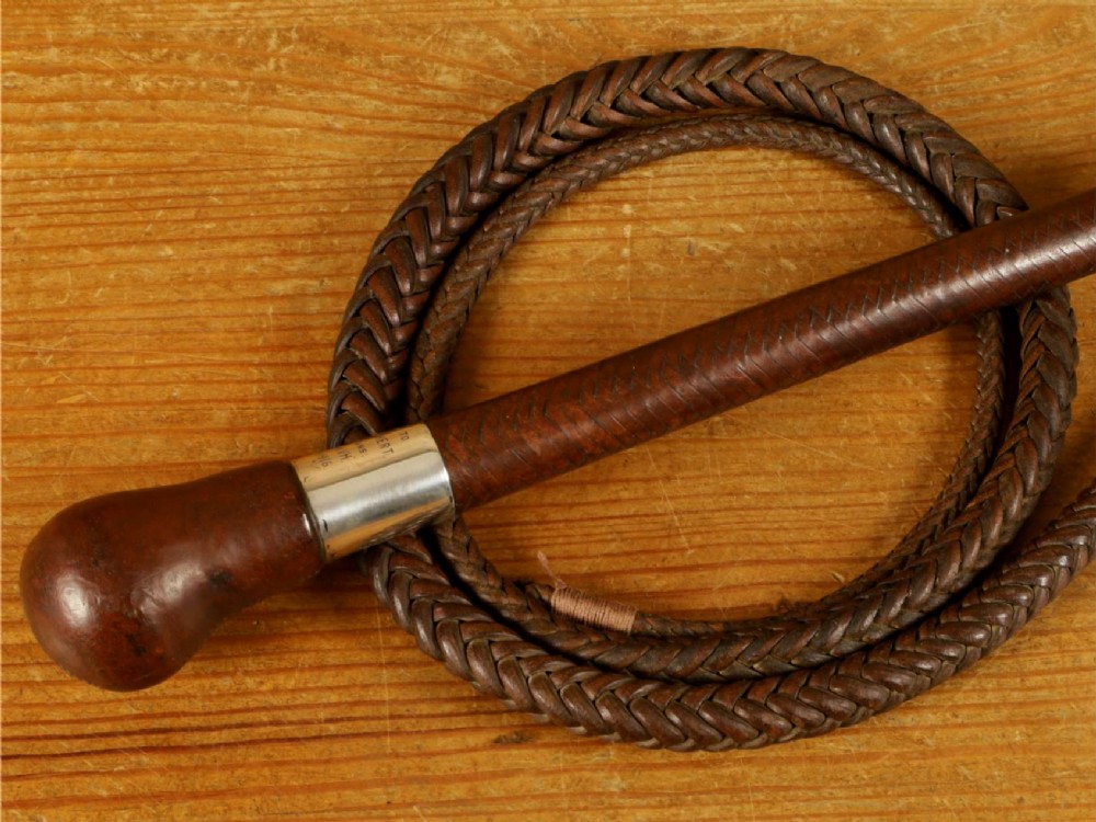 A dark brown braided leather hunting whip with a turned knob handle and an engraved silver collar resting coiled on a wooden surface.