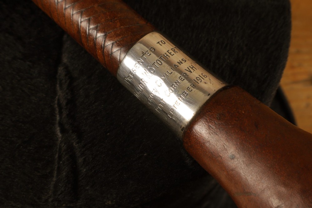 A dark brown braided leather hunting whip with a turned knob handle and an engraved silver collar resting coiled on a wooden surface. View IV
