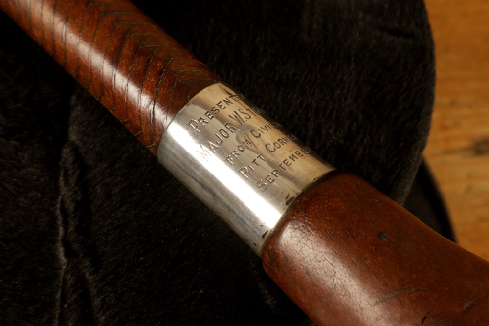 A dark brown braided leather hunting whip with a turned knob handle and an engraved silver collar resting coiled on a wooden surface. View V