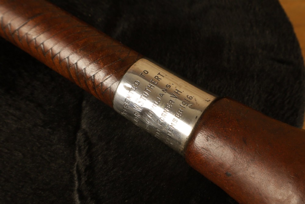 A dark brown braided leather hunting whip with a turned knob handle and an engraved silver collar resting coiled on a wooden surface. View VI