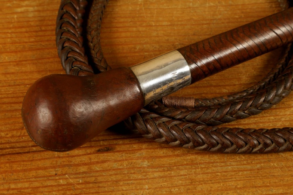 A dark brown braided leather hunting whip with a turned knob handle and an engraved silver collar resting coiled on a wooden surface. View VII