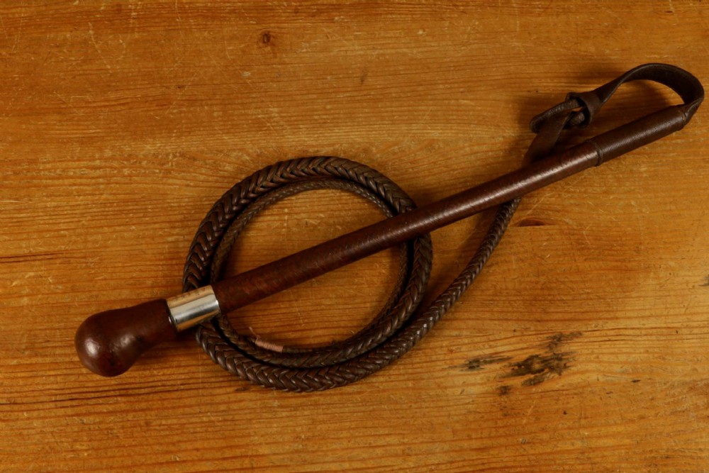 A dark brown braided leather hunting whip with a turned knob handle and an engraved silver collar resting coiled on a wooden surface. View VIII