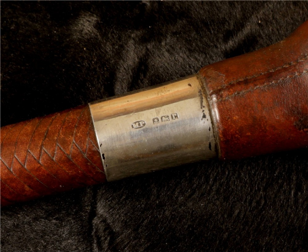 A dark brown braided leather hunting whip with a turned knob handle and an engraved silver collar resting coiled on a wooden surface. View IX