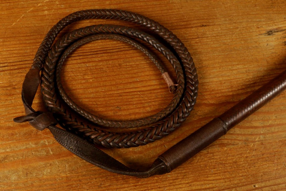 A dark brown braided leather hunting whip with a turned knob handle and an engraved silver collar resting coiled on a wooden surface. View X