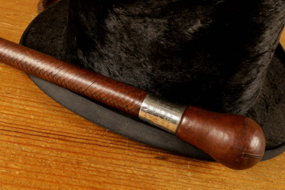 A dark brown braided leather hunting whip with a turned knob handle and an engraved silver collar resting coiled on a wooden surface. View XI