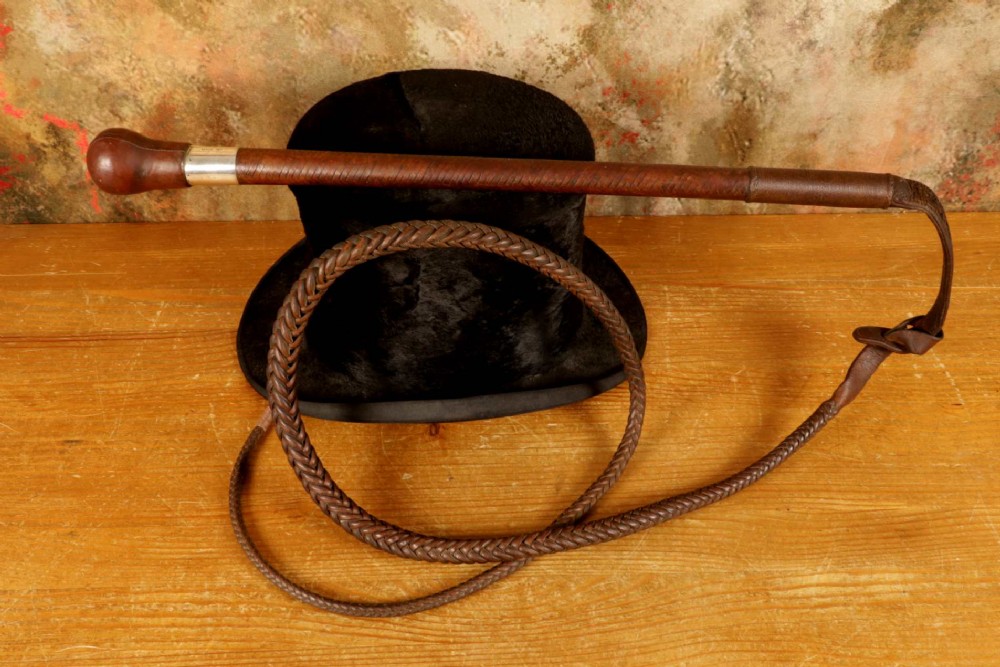 A dark brown braided leather hunting whip with a turned knob handle and an engraved silver collar resting coiled on a wooden surface. View XII