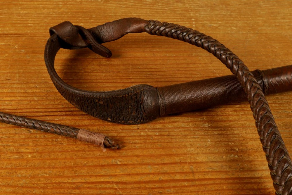 A dark brown braided leather hunting whip with a turned knob handle and an engraved silver collar resting coiled on a wooden surface. View XIII