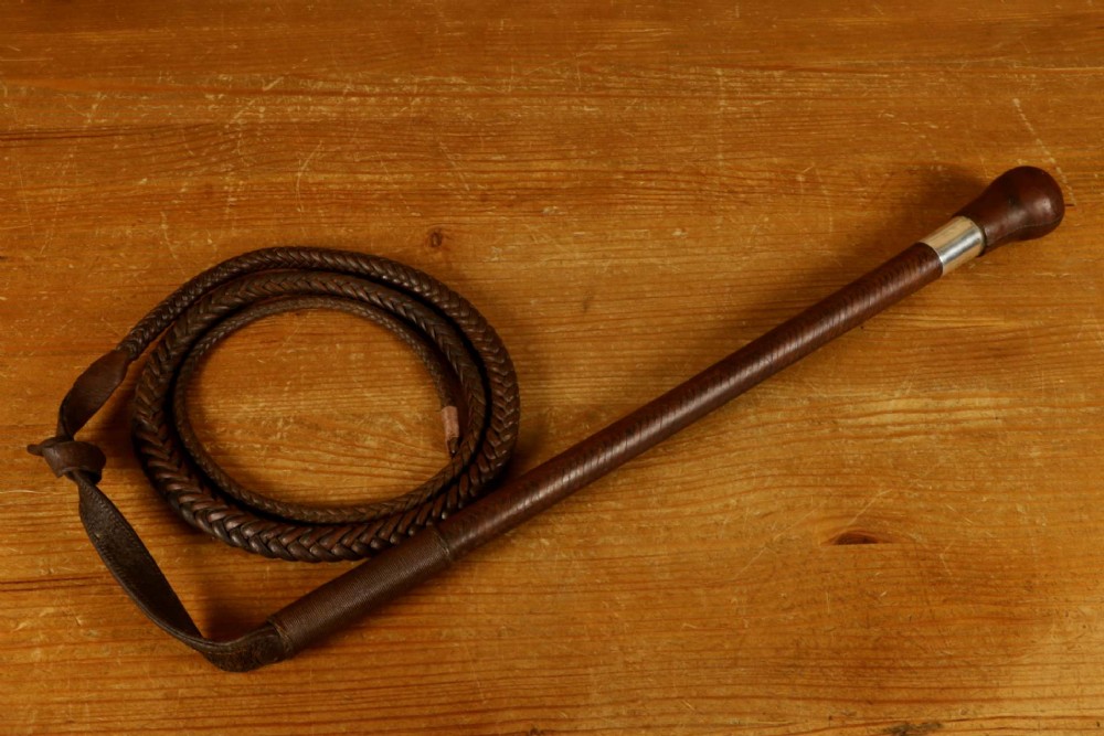 A dark brown braided leather hunting whip with a turned knob handle and an engraved silver collar resting coiled on a wooden surface. View XIV