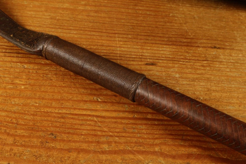 A dark brown braided leather hunting whip with a turned knob handle and an engraved silver collar resting coiled on a wooden surface. View XV
