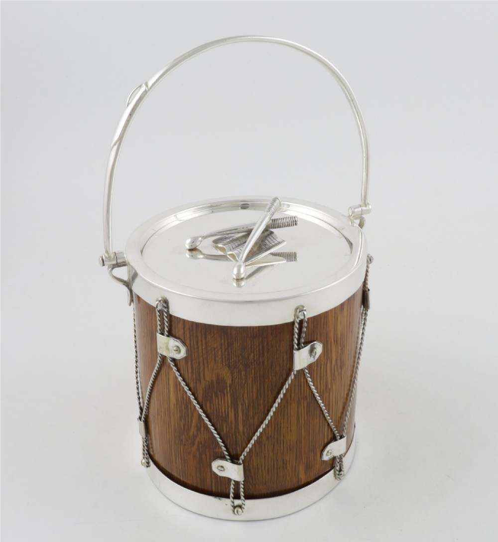 A cylindrical English oak ice pail shaped like a military drum, featuring silver-plated trim, rope-work detailing, a swing handle, and a lid topped with crossed drumsticks. View IV