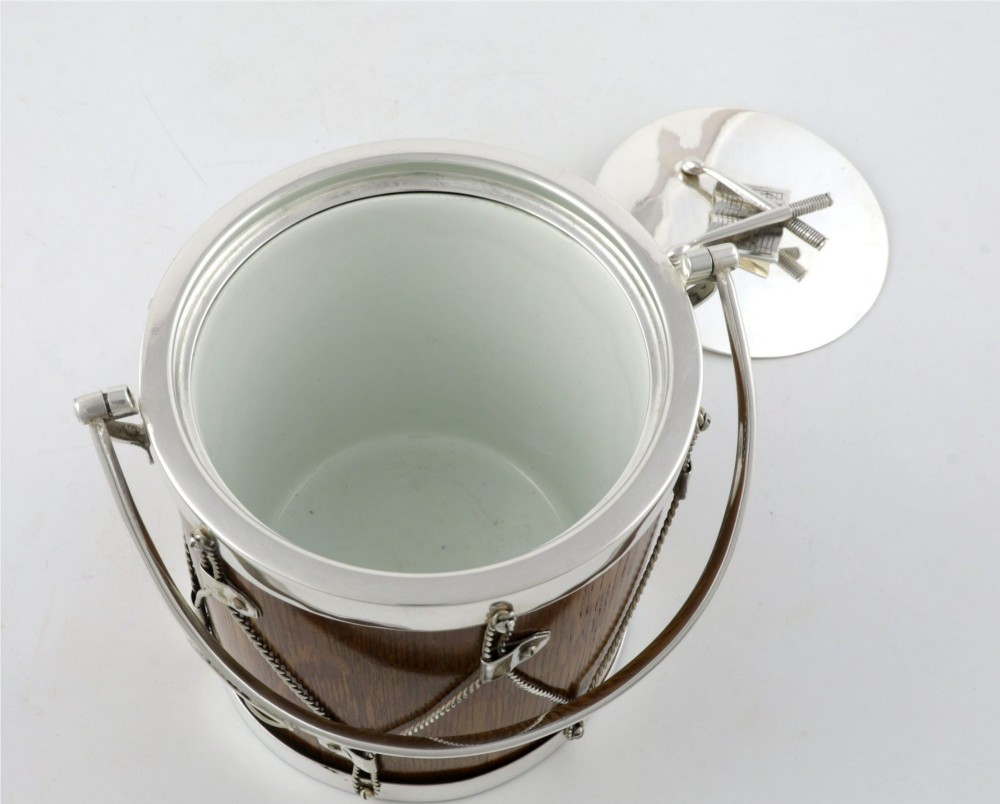 A cylindrical English oak ice pail shaped like a military drum, featuring silver-plated trim, rope-work detailing, a swing handle, and a lid topped with crossed drumsticks. View V