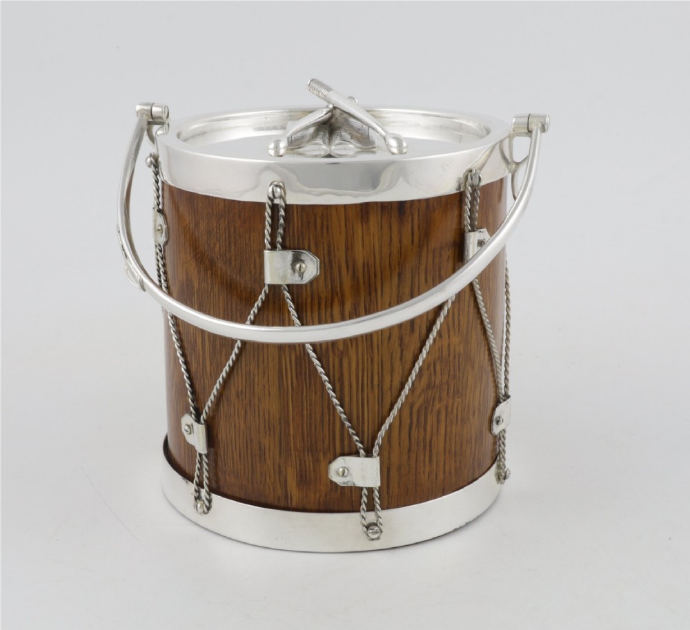 A cylindrical English oak ice pail shaped like a military drum, featuring silver-plated trim, rope-work detailing, a swing handle, and a lid topped with crossed drumsticks. View VI