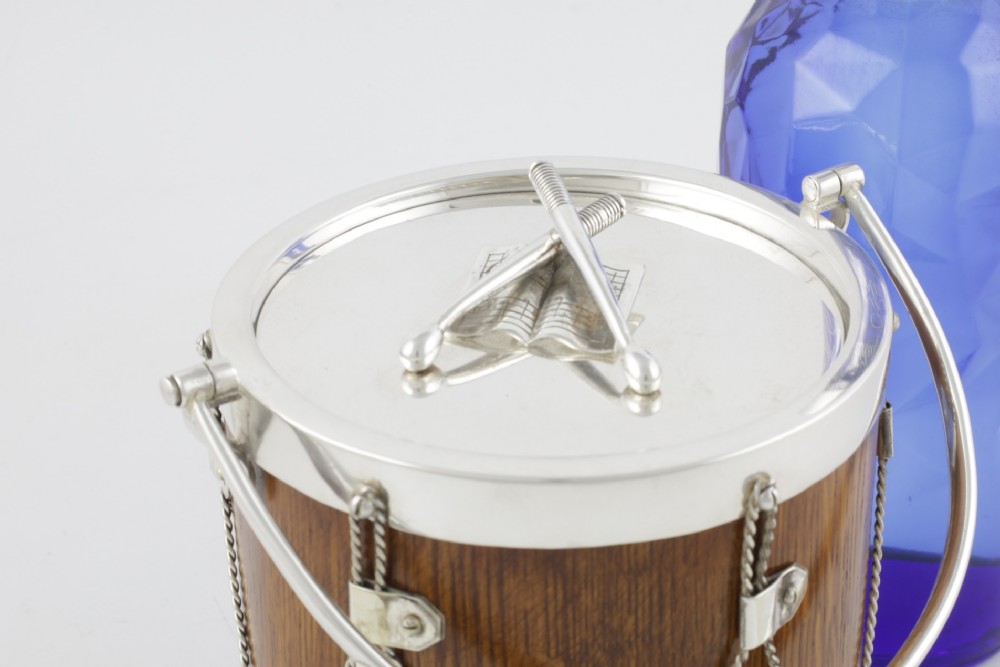 A cylindrical English oak ice pail shaped like a military drum, featuring silver-plated trim, rope-work detailing, a swing handle, and a lid topped with crossed drumsticks. View VII