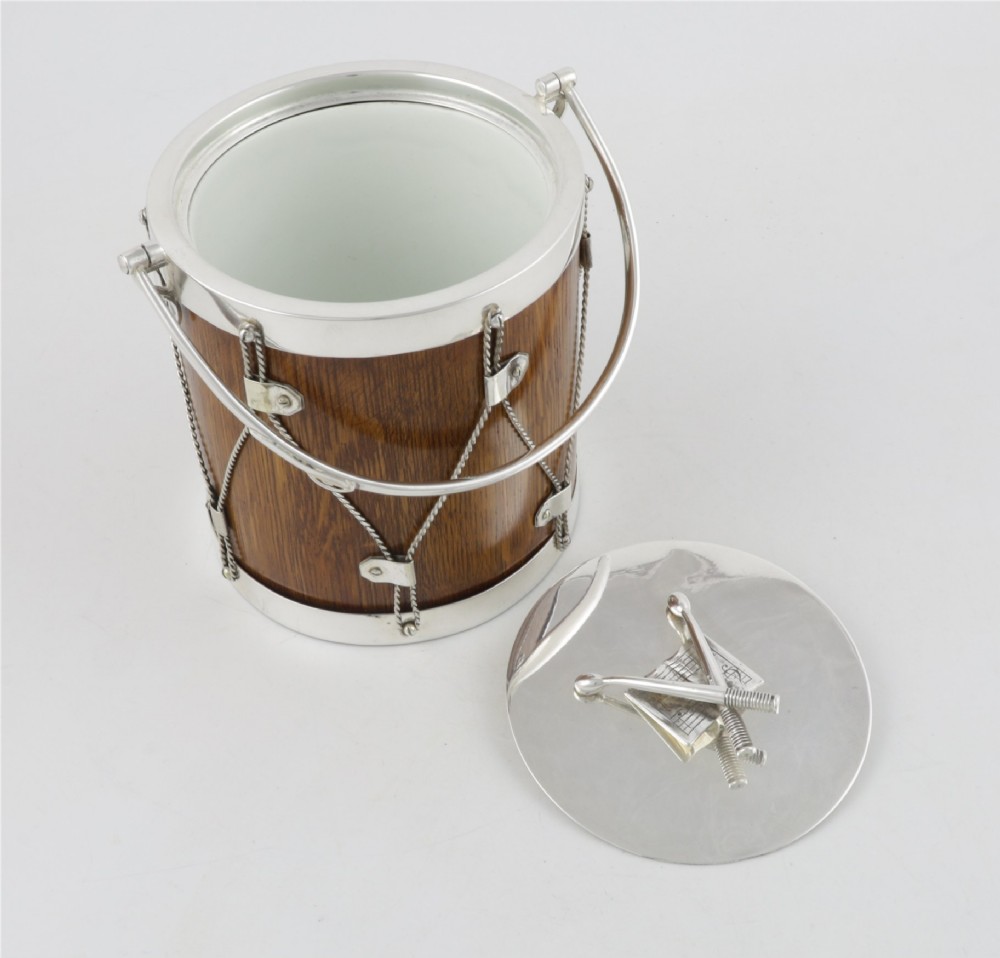 A cylindrical English oak ice pail shaped like a military drum, featuring silver-plated trim, rope-work detailing, a swing handle, and a lid topped with crossed drumsticks. View X