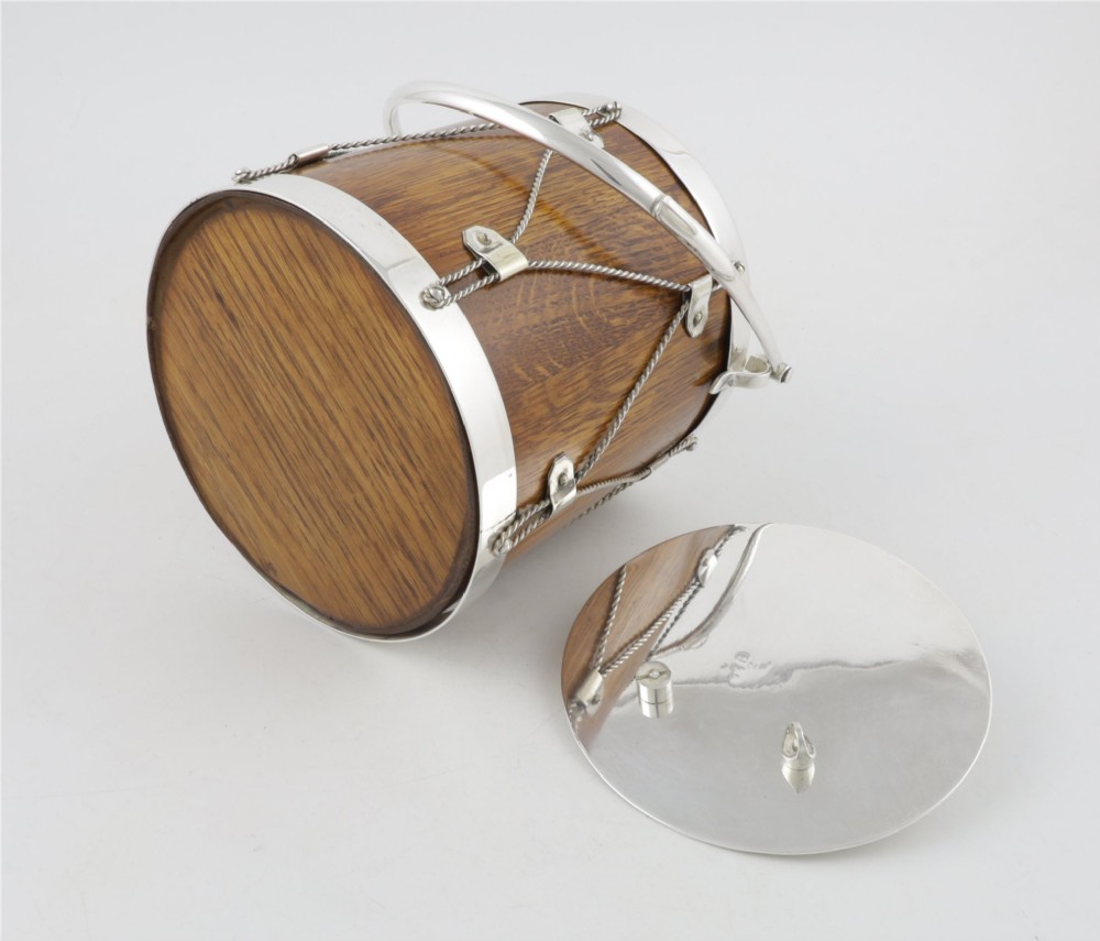 A cylindrical English oak ice pail shaped like a military drum, featuring silver-plated trim, rope-work detailing, a swing handle, and a lid topped with crossed drumsticks. View XII