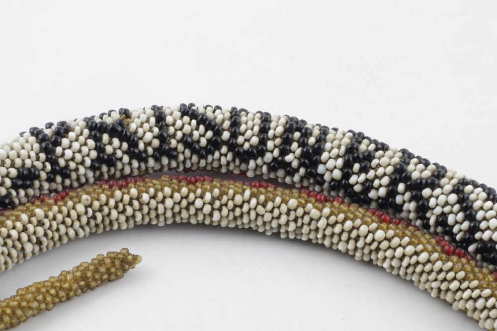 A long, hand-crafted glass beadwork snake with white, olive green, and red geometric patterns coiled into a spiral, featuring text reading TURKISH PRISONERS 1918 woven into its body. View IV