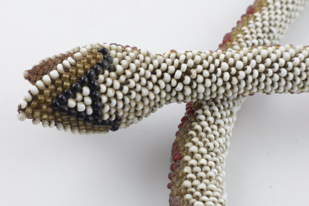 A long, hand-crafted glass beadwork snake with white, olive green, and red geometric patterns coiled into a spiral, featuring text reading TURKISH PRISONERS 1918 woven into its body. View VI