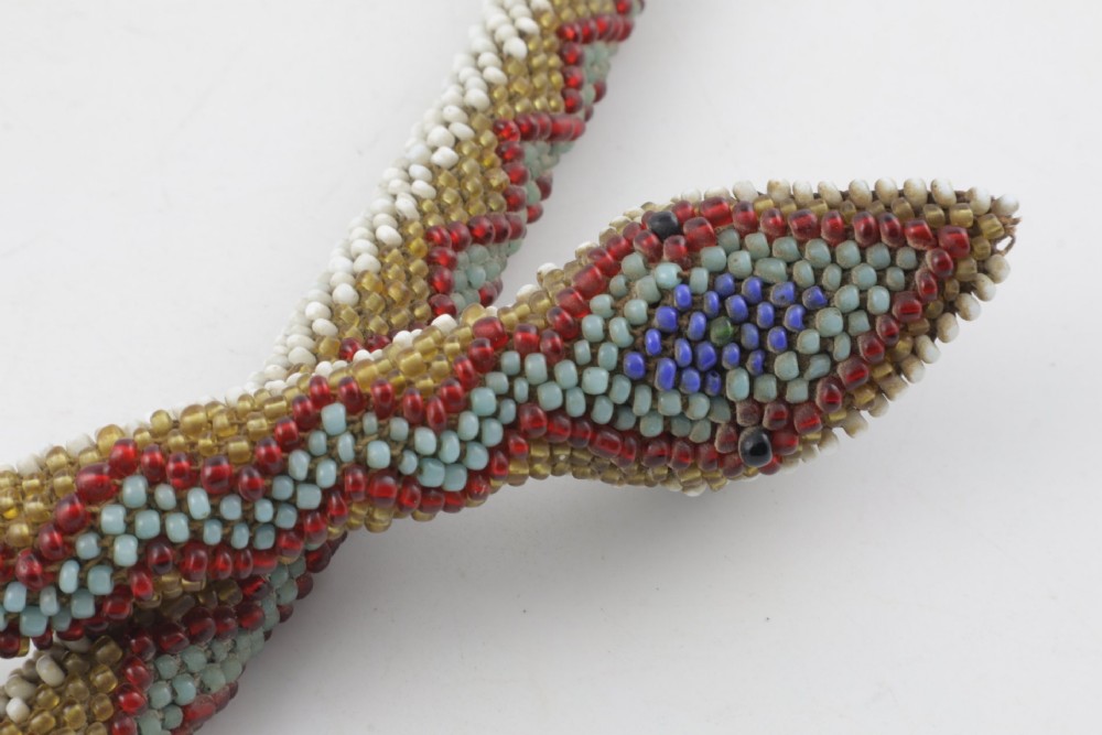 A long, hand-crafted glass beadwork snake with white, olive green, and red geometric patterns coiled into a spiral, featuring text reading TURKISH PRISONERS 1918 woven into its body. View VII