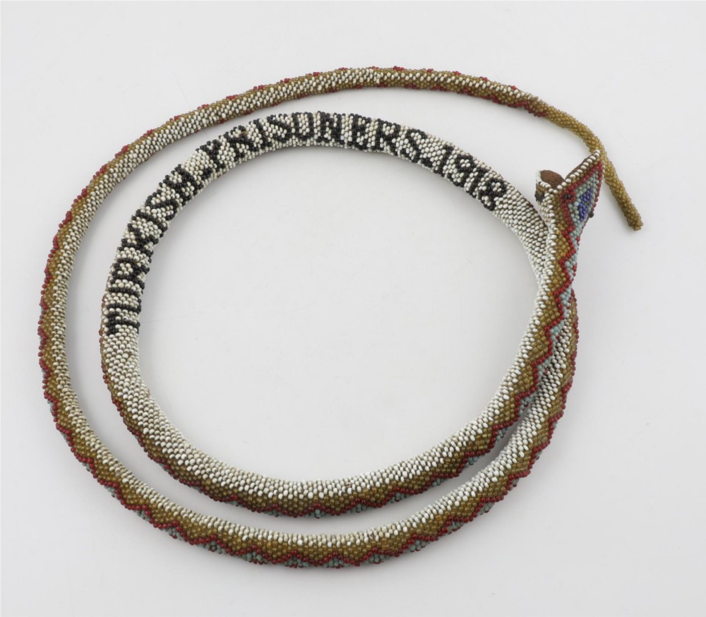 A long, hand-crafted glass beadwork snake with white, olive green, and red geometric patterns coiled into a spiral, featuring text reading TURKISH PRISONERS 1918 woven into its body. View VIII