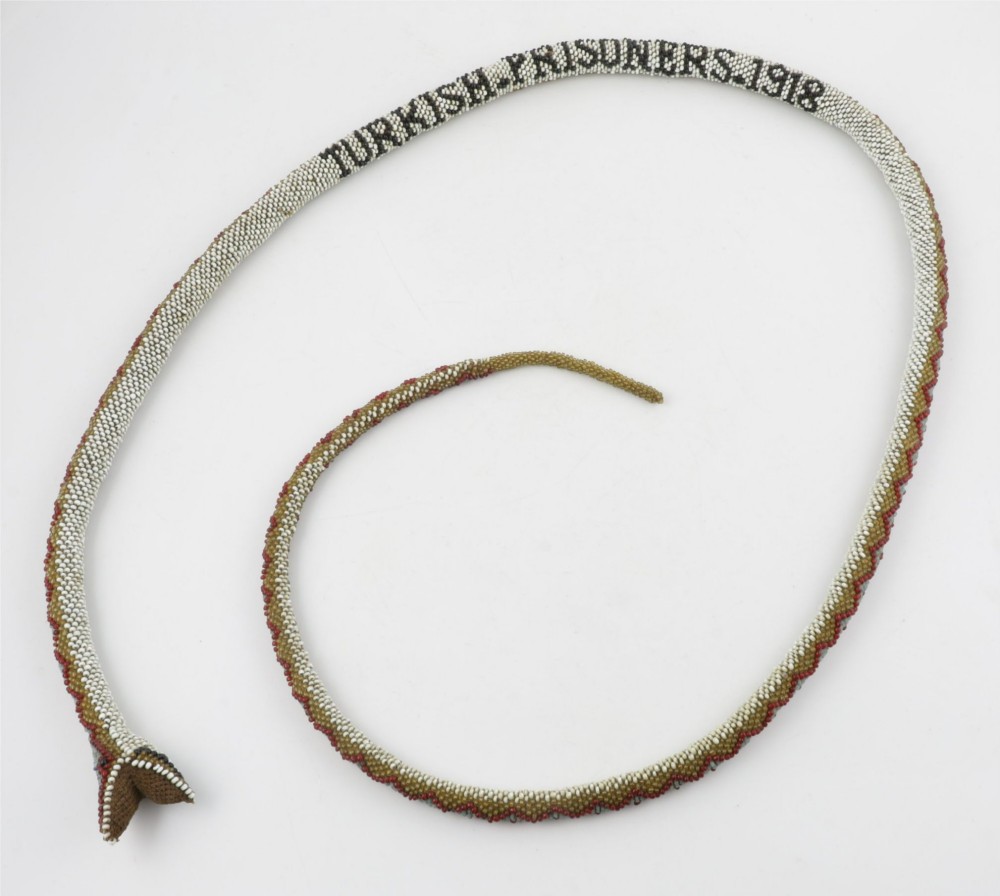 A long, hand-crafted glass beadwork snake with white, olive green, and red geometric patterns coiled into a spiral, featuring text reading TURKISH PRISONERS 1918 woven into its body. View IX