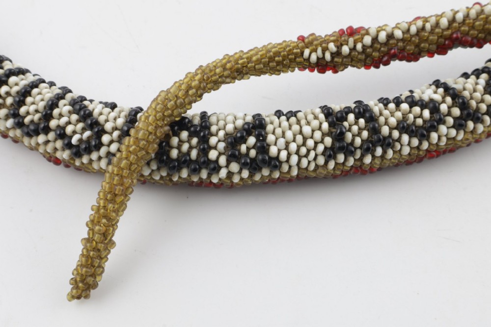 A long, hand-crafted glass beadwork snake with white, olive green, and red geometric patterns coiled into a spiral, featuring text reading TURKISH PRISONERS 1918 woven into its body. View X