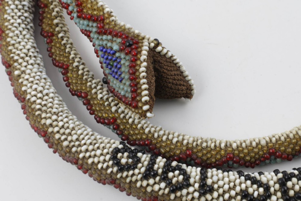 A long, hand-crafted glass beadwork snake with white, olive green, and red geometric patterns coiled into a spiral, featuring text reading TURKISH PRISONERS 1918 woven into its body. View XIII