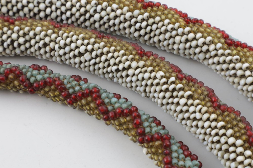 A long, hand-crafted glass beadwork snake with white, olive green, and red geometric patterns coiled into a spiral, featuring text reading TURKISH PRISONERS 1918 woven into its body. View XIV