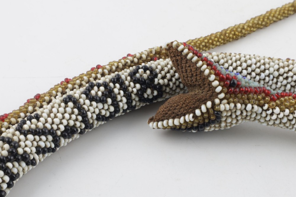A long, hand-crafted glass beadwork snake with white, olive green, and red geometric patterns coiled into a spiral, featuring text reading TURKISH PRISONERS 1918 woven into its body. View XV