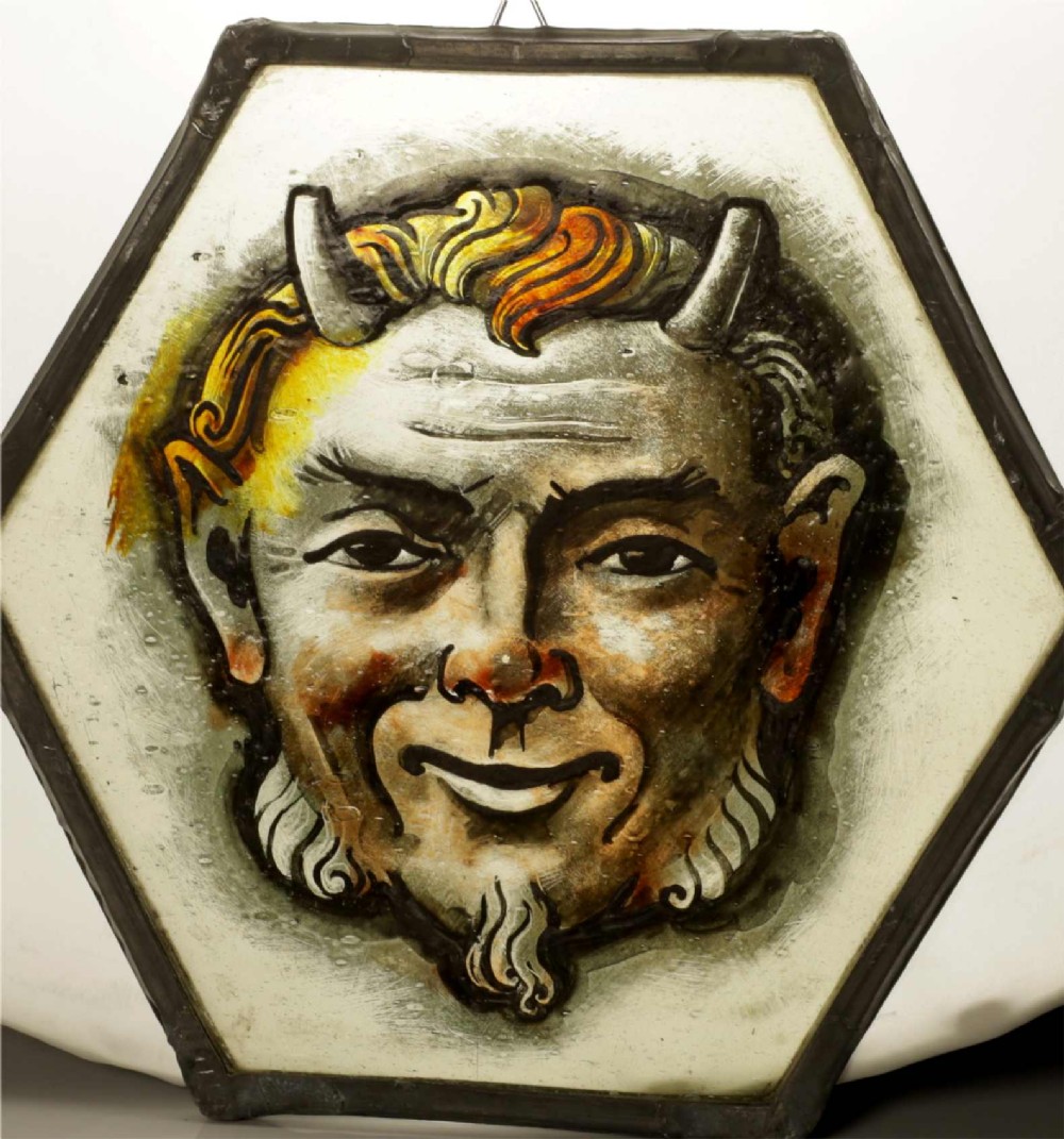 A hexagonal leaded stained glass panel featuring a hand-painted depiction of a horned devil head in grey, black, and amber tones.