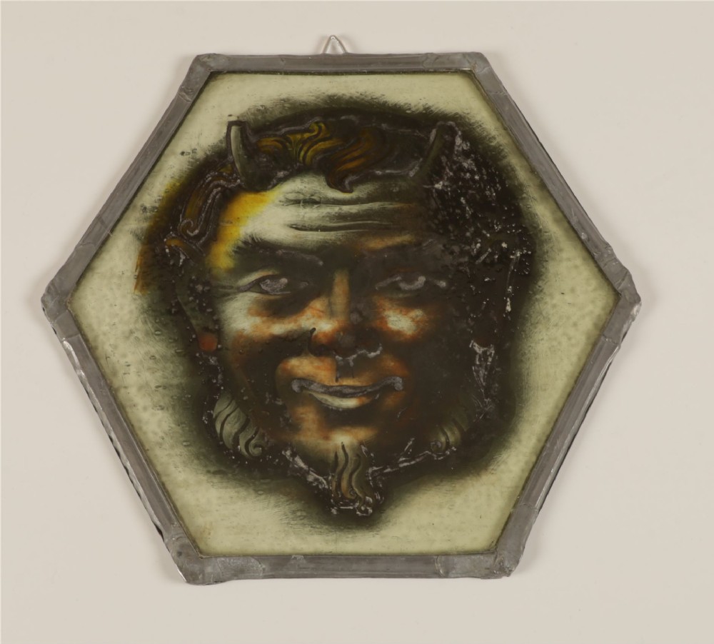 A hexagonal leaded stained glass panel featuring a hand-painted depiction of a horned devil head in grey, black, and amber tones. View IV