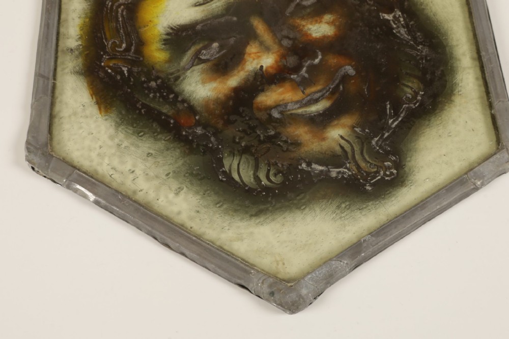 A hexagonal leaded stained glass panel featuring a hand-painted depiction of a horned devil head in grey, black, and amber tones. View V