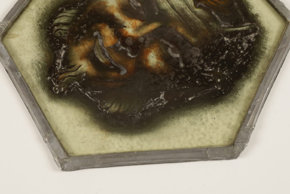 A hexagonal leaded stained glass panel featuring a hand-painted depiction of a horned devil head in grey, black, and amber tones. View VII