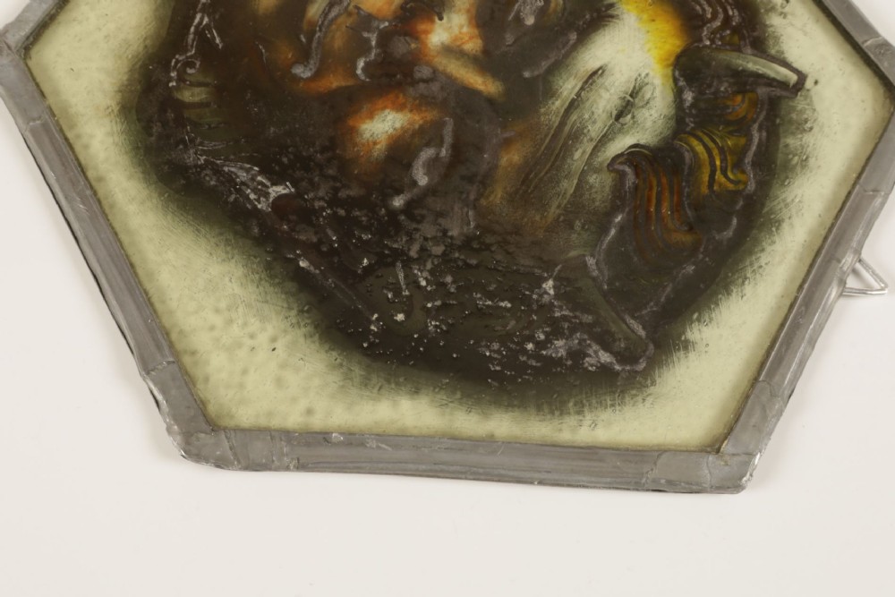 A hexagonal leaded stained glass panel featuring a hand-painted depiction of a horned devil head in grey, black, and amber tones. View VIII