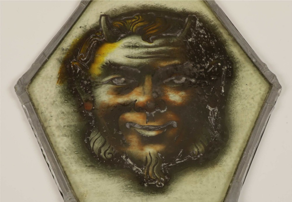 A hexagonal leaded stained glass panel featuring a hand-painted depiction of a horned devil head in grey, black, and amber tones. View IX