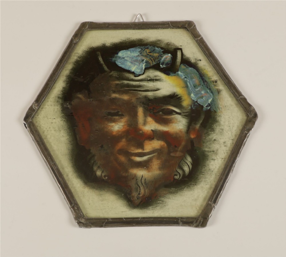 A hexagonal leaded stained glass panel featuring a hand-painted depiction of a horned devil head in grey, black, and amber tones. View XII