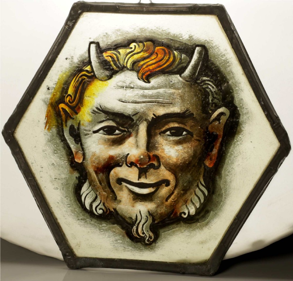 A hexagonal leaded stained glass panel featuring a hand-painted depiction of a horned devil head in grey, black, and amber tones. View XIV