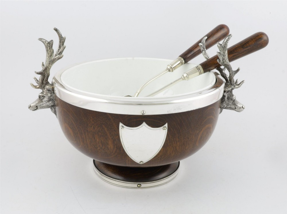 A turned dark oak bowl featuring a silver-plated rim, cast stag head handles, a central vacant silver shield, and matching serving utensils.