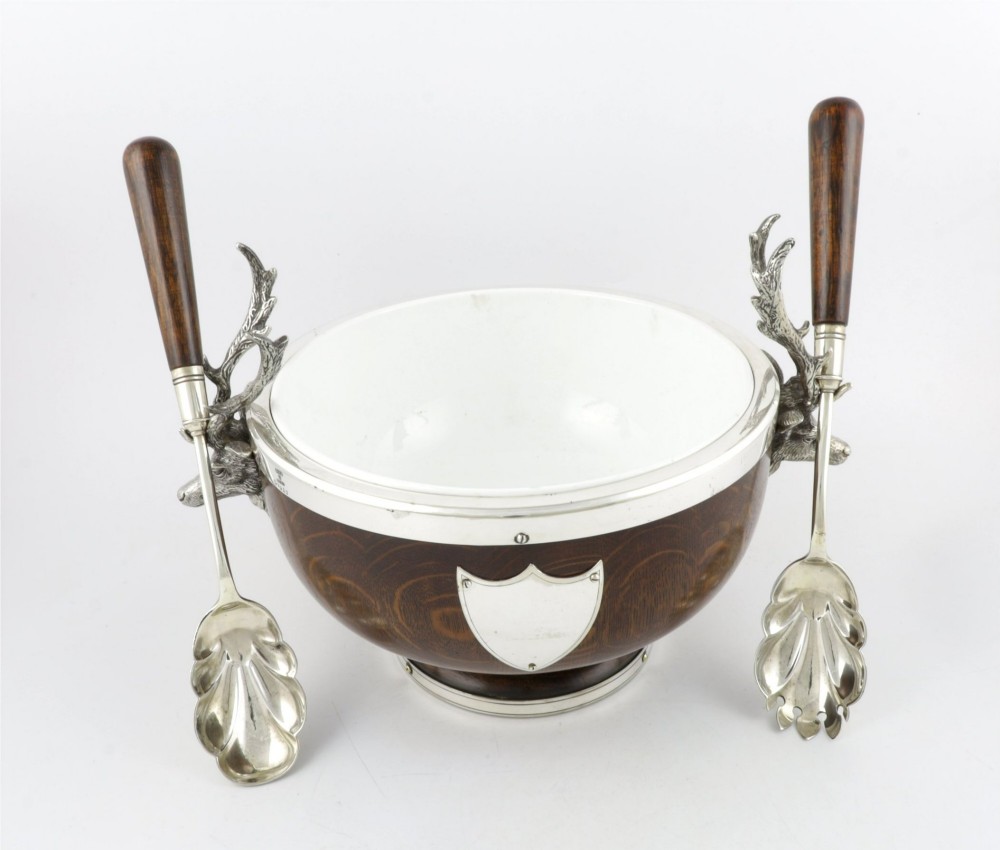 A turned dark oak bowl featuring a silver-plated rim, cast stag head handles, a central vacant silver shield, and matching serving utensils. View V