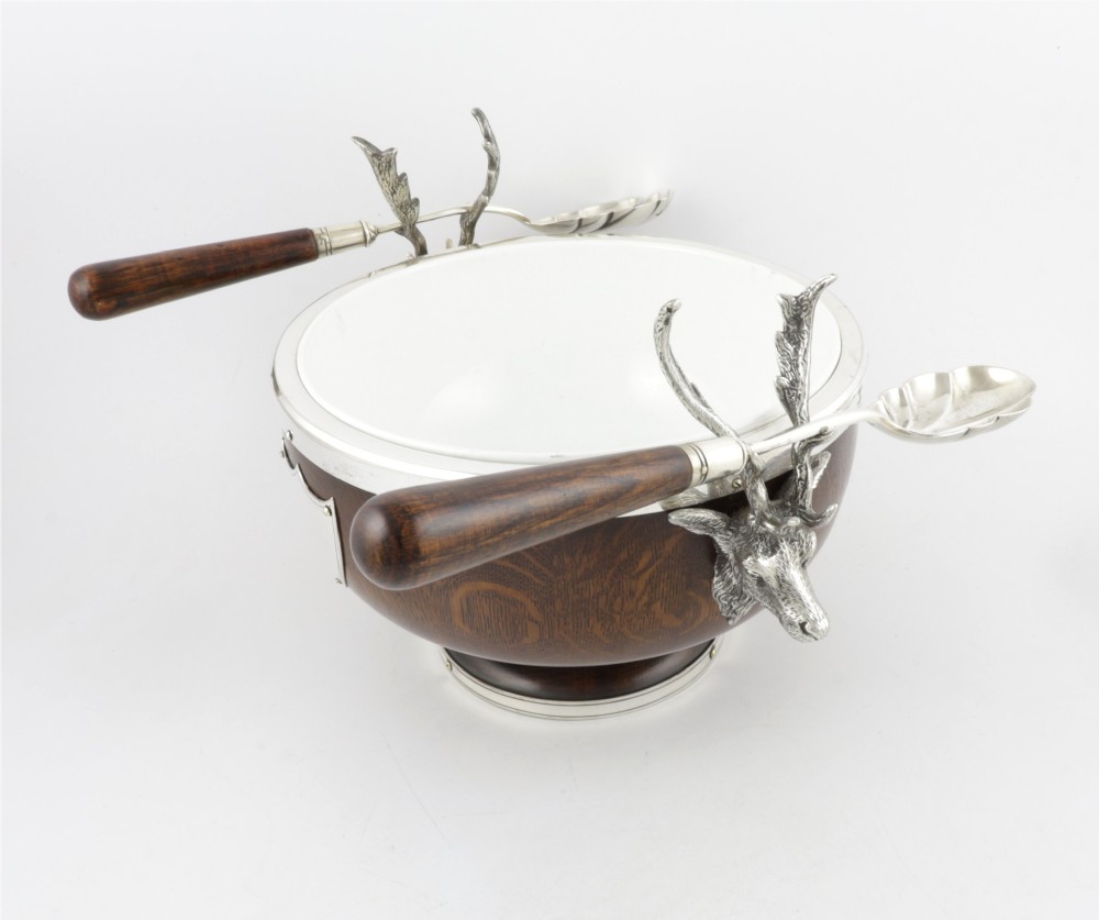 A turned dark oak bowl featuring a silver-plated rim, cast stag head handles, a central vacant silver shield, and matching serving utensils. View VI