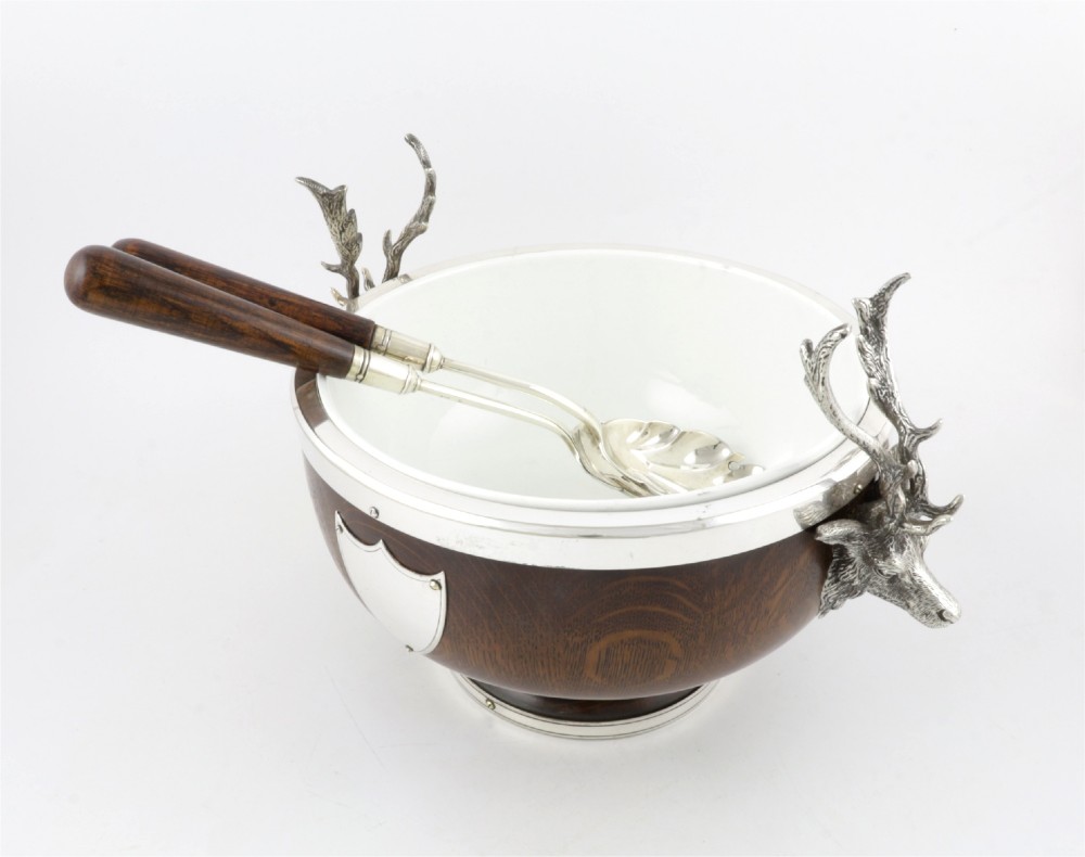 A turned dark oak bowl featuring a silver-plated rim, cast stag head handles, a central vacant silver shield, and matching serving utensils. View VII