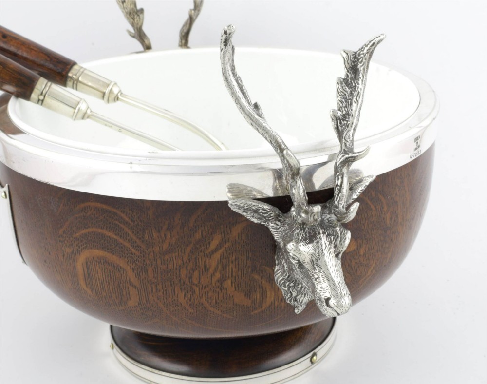 A turned dark oak bowl featuring a silver-plated rim, cast stag head handles, a central vacant silver shield, and matching serving utensils. View IX