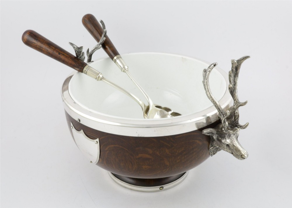 A turned dark oak bowl featuring a silver-plated rim, cast stag head handles, a central vacant silver shield, and matching serving utensils. View X